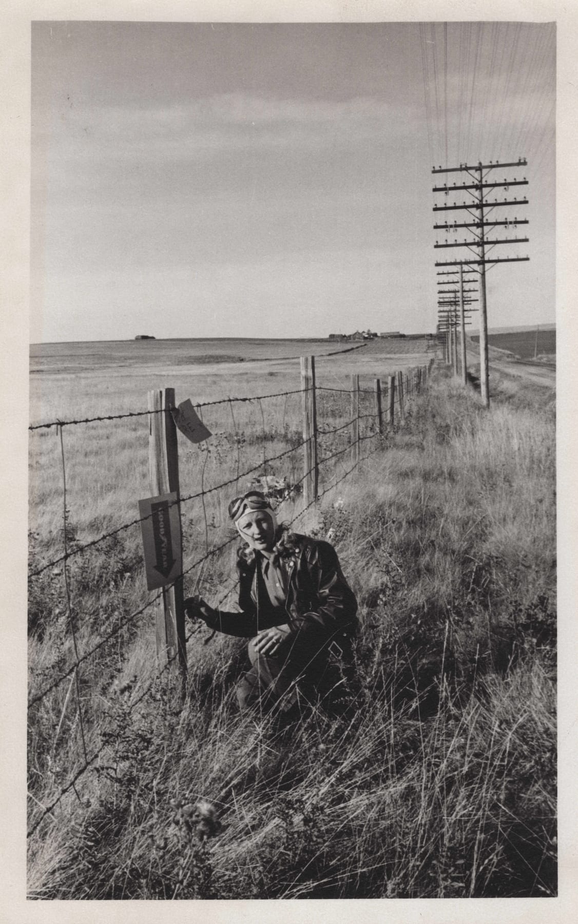 A rider in leather jacket and goggles crouched at a barbed-wire fence beside a clue and a Goodyear sign