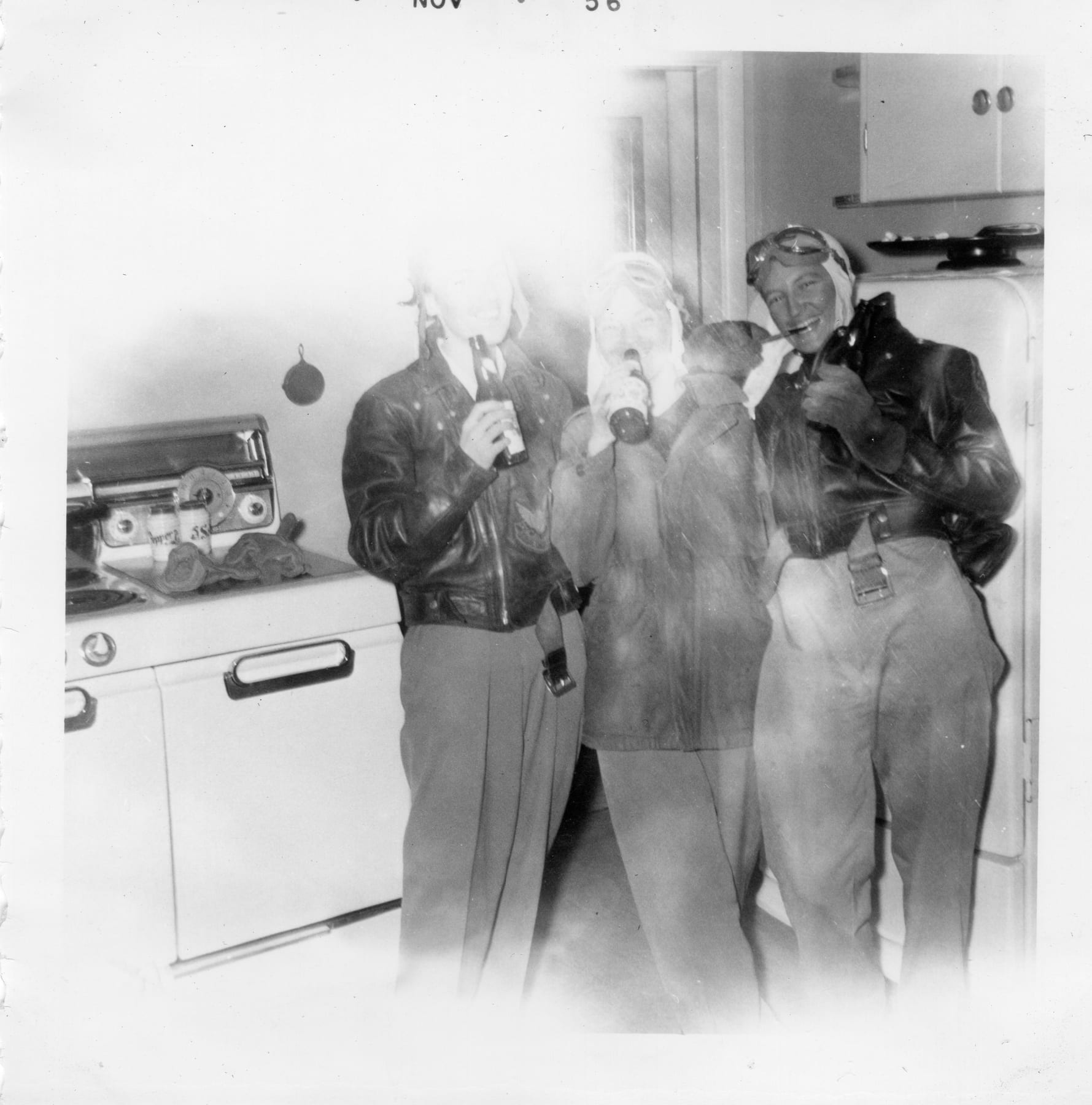 Ace-Hy members in leather jackets, caps and goggles with drinks in hand in a 1950s kitchen